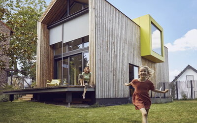 Kind läuft fröhlich von einem sehr ökologisch-modern aussehendem Haus weg, auf dessen Terasse eine Frau sitzt